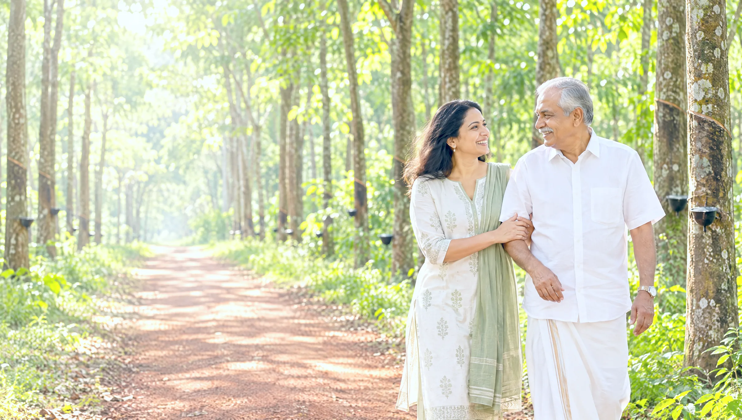A woman walking arm in arm with her father along a sunlit lane through a rubber plantation near Kottayam